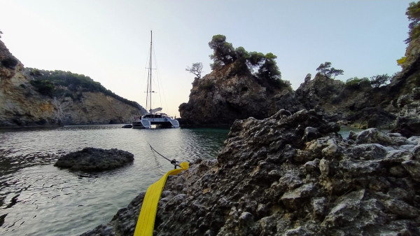 Skippertraining bei Korfu Segeln Yacht mit Anker voraus und langer Landleine um einen Felsen gewunden in malerischer Bucht mit Klippen